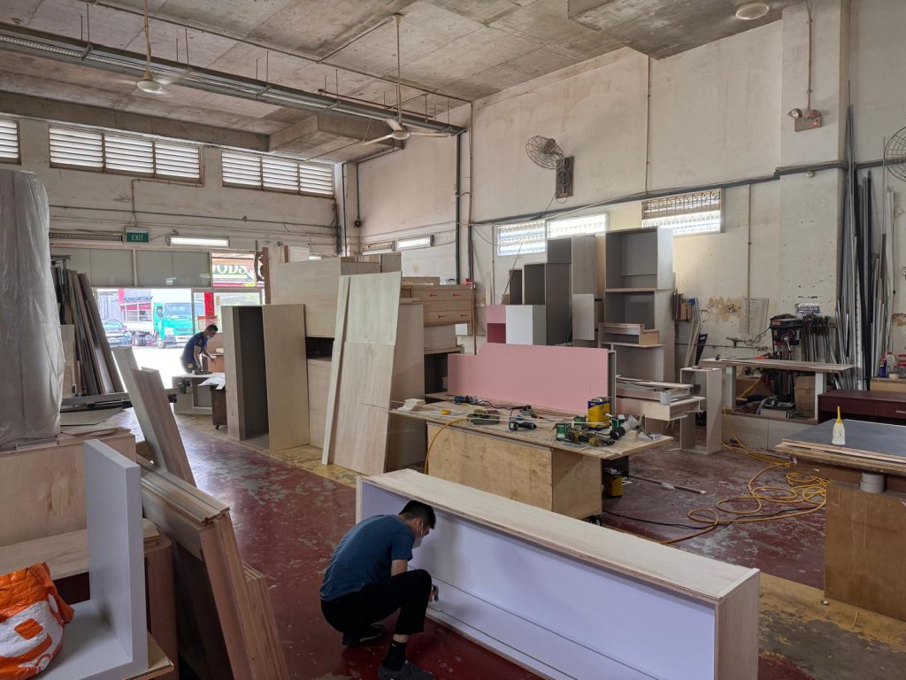 Carpenters assembling custom cabinetry among workbenches and stacked panels in Innovation Workshop’s workshop